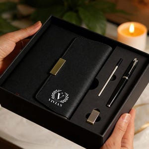 Può includere: Un set regalo nero contenente un taccuino, un portafoglio, una penna e una matita. Il portafoglio è impresso con il nome "VIVIAN" e un'iniziale "V". Il set include un accento metallico color oro sul portafoglio e una clip metallica color argento.