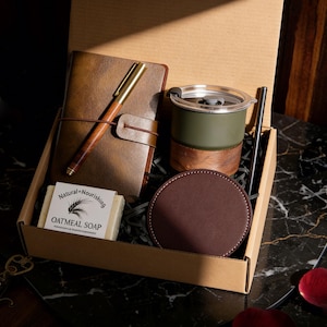 Könnte beinhalten: Eine Geschenkbox mit einem braunen Lederjournal, einem Holzstift, einem grün- und holzfarbenen Reisebecher, einem braunen Lederuntersetzer, einem Stück Haferflocken-Seife und einem schwarzen Metallstrohhalm. Die Seife ist mit "Natural Nourishing Oatmeal Soap" beschriftet.