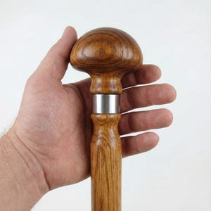 Può includere: Bastone da passeggio in legno con manico a forma di fungo. Il manico è marrone chiaro, con una fascia in metallo argentato. L'asta è anch'essa marrone chiaro. Il bastone è tenuto in mano su sfondo bianco.