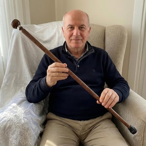 May include: A dark brown wooden walking stick with a rounded, dark brown top and a silver accent. The stick has a rubber ferrule and is held by a person sitting in a chair.