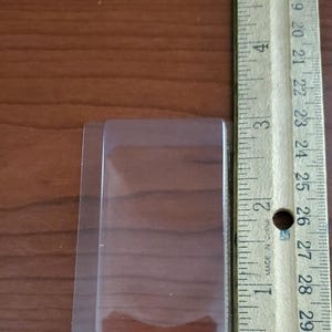 May include: A clear plastic blister pack with a rectangular shape, next to a wooden ruler. The ruler shows measurements in inches, with the blister pack measuring approximately 3 inches in length.