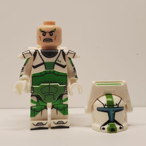 May include: A plastic minifigure featuring a white and green armored torso and legs with black detailing. The figure has a flesh-toned head with a stern expression and green markings above the eyes. A matching white and green helmet sits nearby.