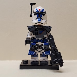 May include: A white, blue, and grey plastic toy soldier minifigure wearing a helmet with a black antenna. The figure features detailed armor printing, a blue fabric-style waist cape, and holds a large black plastic blaster weapon in one hand.