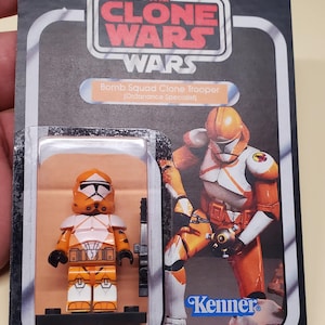 May include: A Star Wars The Clone Wars Bomb Squad Clone Trooper action figure in original blister card packaging. The figure is orange and white with a matching helmet and armor. The card features the Kenner logo and character artwork.