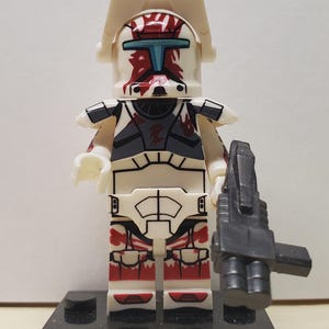 May include: A white plastic toy soldier minifigure featuring red markings on the helmet and legs, a dark gray chest plate, and a white visor attachment. The figure holds a black plastic blaster weapon and stands on a rectangular black base.
