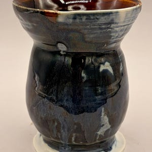May include: A handmade ceramic vase with a unique shape and a glossy finish. The vase features a dark blue and brown color scheme with white accents at the base. The interior is a rich brown color.
