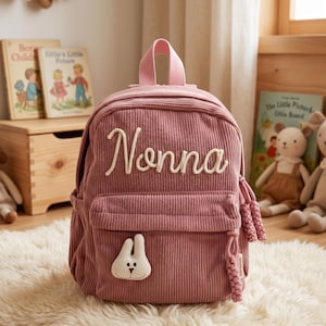 Peut inclure: Un sac à dos en velours côtelé rose poudré avec le nom "Nonna" brodé en cursive blanche. Le sac à dos présente un petit appliqué de lapin et des pompons tressés. Une poignée supérieure et des bretelles sont visibles.