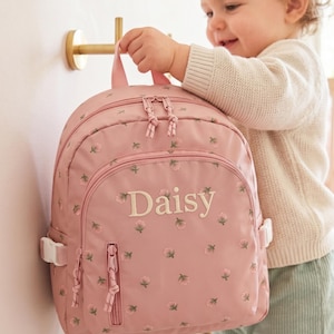 May include: Pink backpack with floral embroidery and the name "Daisy" embroidered on the front. The backpack has a top handle, two zippered compartments, and adjustable straps. The backpack is designed for children.