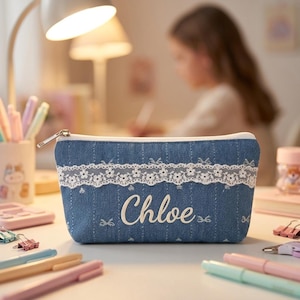 May include: A denim pencil case with a white lace trim and the name "Chloe" embroidered in cursive. The case has a zipper closure and is surrounded by pens and other stationery items.