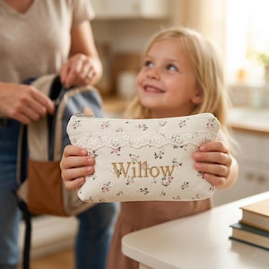 Puede incluir: Un estuche crema con un estampado floral y el nombre "Willow" bordado en dorado. El estuche tiene un ribete de encaje y lo sostiene una niña. Una mochila está en el fondo.