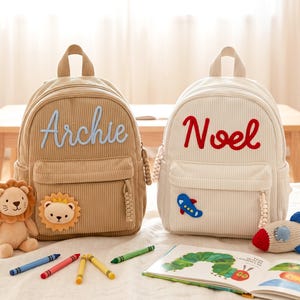 May include: Two corduroy backpacks, one tan with the name Archie in light blue and a lion patch, and one cream with the name Noel in red and a blue airplane patch. Both feature a top handle, a front zippered pocket, and a braided zipper pull.
