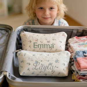 May include: Two floral print cosmetic bags with lace trim, one embroidered with "Emma" in green and the other with "Layla" in silver, are inside an open suitcase. The bags have a zipper closure and are filled with items.