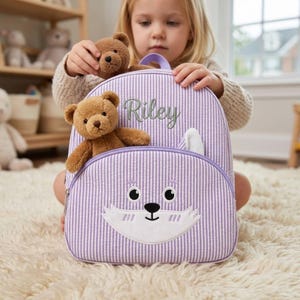 May include: A light purple and white striped backpack with a fox face design on the front. The backpack has a top handle and a front pocket. The name "Riley" is embroidered on the front. Two small brown teddy bears are attached to the backpack.