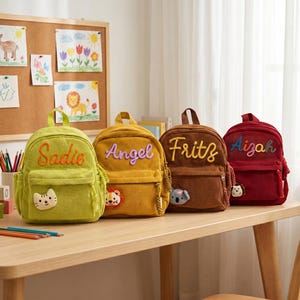 Peut inclure: Quatre sacs à dos en velours côtelé vert, jaune, marron et rouge, chacun avec un nom brodé sur le devant. Le sac vert a un patch chat, le jaune a un lion, le marron a un koala et le rouge a un hérisson.