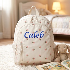 Puede incluir: Una mochila color crema con un estampado floral de rosas rosas. El nombre "Caleb" está bordado en azul en la parte delantera. La mochila tiene un asa superior, un bolsillo delantero y bolsillos laterales. Un conejito de peluche y un libro están al lado.