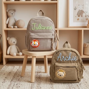 Peut inclure: Deux sacs à dos en velours côtelé, un beige et un marron, avec des noms brodés et des écussons de lion. Le sac à dos beige porte "Julianne" en écriture verte, et le sac à dos marron porte "Archie" en écriture bleue. Les deux ont une poche avant et des détails latéraux tressés.
