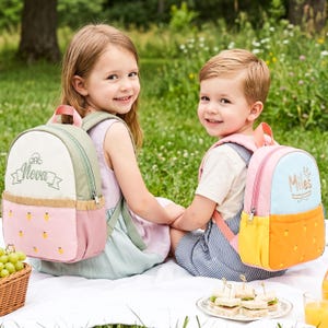 May include: Two small backpacks with embroidered names. The left backpack is sage green, cream, and pink with a mushroom icon and the name Nova. The right backpack is light blue, pink, and yellow with a bird icon and the name Miles.