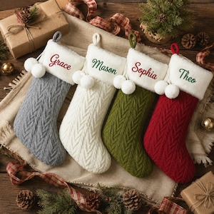 May include: Four cable-knit Christmas stockings in gray, white, olive green, and red. Each stocking features a white plush cuff embroidered with the names Grace, Mason, Sophia, and Theo, and is decorated with two white pom-poms on a hanging loop.