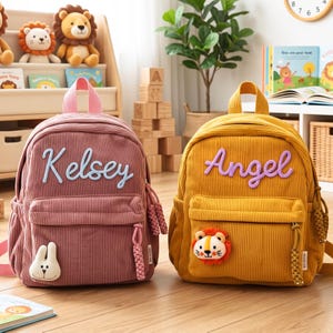 May include: Two corduroy backpacks, one dusty rose and one mustard yellow. The rose backpack features the name Kelsey in light blue script and a white rabbit patch. The yellow backpack features the name Angel in purple script and a lion patch.