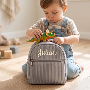 Könnte beinhalten: Ein grauer Cord-Rucksack mit dem Namen "Julian" in Weiß gestickt. Ein grünes und orangefarbenes Spielzeug-Dinosaurier sitzt oben auf dem Rucksack. Der Rucksack hat eine Reißverschluss-Vordertasche und einen oberen Griff.