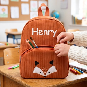 May include: A burnt orange corduroy backpack with the name "Henry" in white. The backpack has a fox face design on the front pocket. Pencils and a paintbrush are visible in the front pocket. A child is reaching into the pocket.