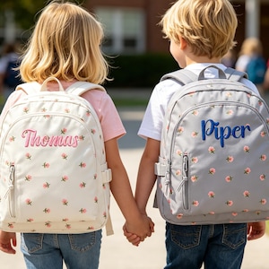 May include: Two children holding hands, each wearing a backpack. One backpack is off-white with pink floral accents and the name "Thomas" embroidered in pink. The other is gray with pink floral accents and the name "Piper" embroidered in blue.