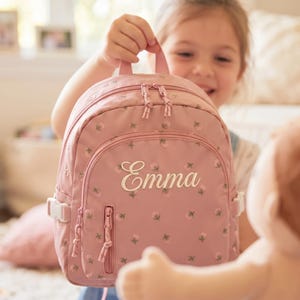 以下が含まれることがあります： 前面に「Emma」と刺繍された、花柄のピンク色のバックパック。 バックパックには、上部のハンドル、2つのジッパー付きコンパートメント、調節可能なストラップが付いています。 バッグは人が持っています。
