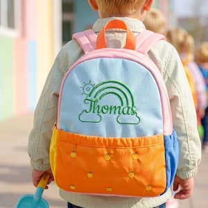 May include: A multi-colored backpack featuring a light blue upper section with an embroidered green rainbow, a sun, and the name Thomas. The lower section is orange with small embroidered fruit motifs, complemented by pink straps and a blue side pocket.