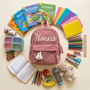 Peut inclure: Un sac à dos en velours côtelé rose avec le nom "Nonna" brodé dessus, entouré de fournitures scolaires. Comprend des livres, des cahiers, des crayons, des crayons de couleur, une boîte à lunch, une bouteille d'eau, un ours en peluche et des fruits.