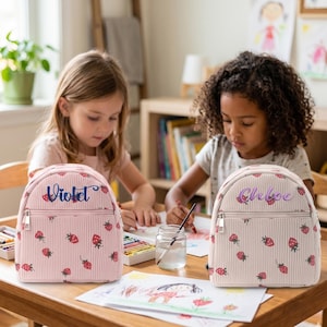 Puede incluir: Dos pequeñas mochilas con estampados de fresas y nombres personalizados. Una es rosa claro con el nombre "Violet" en escritura azul, y la otra es blanquecina con el nombre "Chloe" en escritura morada. Ambas tienen un patrón de rayas verticales y un bolsillo frontal con cremallera.