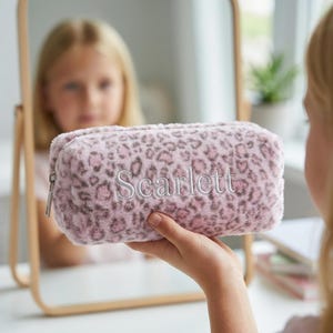 May include: A pink and gray leopard print pencil case with the name "Scarlett" embroidered in white. The rectangular case has a zipper closure and is made of a soft, plush material. The case is held up in front of a mirror.
