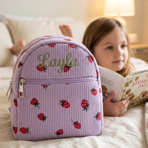 Könnte beinhalten: Ein lavendelfarbener Cord-Rucksack mit Reißverschluss und einem sich wiederholenden Erdbeermuster. Der Name "Layla" ist in grüner Schrift aufgestickt. Der Rucksack ist für Kinder.