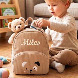 May include: Tan corduroy backpack with a dog face design on the front, featuring the name "Miles" in cursive. A teddy bear and a small sock are in the backpack. The backpack has a top handle and adjustable straps.