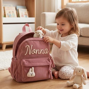 May include: A dusty rose corduroy backpack featuring the name Nonna embroidered in white script on the front pocket. The bag includes a small white plush sheep toy peeking out from the top and a white plush rabbit patch on the lower pocket.