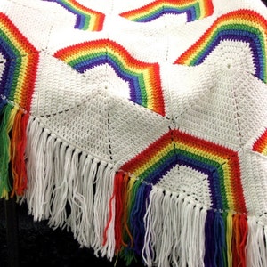 May include: A white crocheted blanket with rainbow stripes and fringe. The blanket is made of a soft, textured yarn and has a delicate, handmade look.