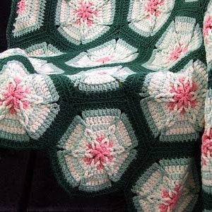 May include: A handmade crochet blanket featuring a floral pattern. The blanket is composed of dark green, white, light blue, and pink hexagonal motifs. The floral design is centered with pink and surrounded by white and light blue petals.