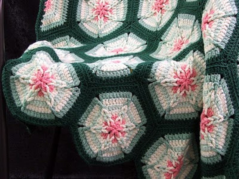 May include: A handmade crochet blanket featuring a floral pattern. The blanket is composed of dark green, white, light blue, and pink hexagonal motifs. The floral design is centered with pink and surrounded by white and light blue petals.