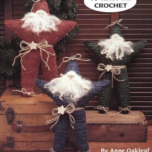 May include: Three knitted and crocheted star-shaped ornaments, each depicting a Santa Claus figure with a white beard and a star on their belly. The ornaments are red, green, and blue. The title "To Knit and Crochet" is displayed above the ornaments, and the text "By Anne Oakleaf" is displayed below the ornaments.