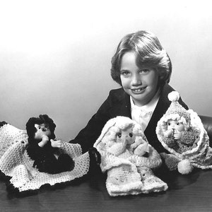 Könnte beinhalten: Schwarz-Weiß-Fotografie einer lächelnden jungen Person mit drei handgefertigten Häkelpuppen. Die Puppen haben bemalte Gesichter und sind in Häkeloutfits gekleidet, darunter Hüte und Decken. Die Puppen befinden sich auf einer dunklen Oberfläche.