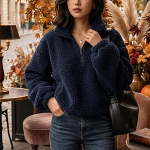 Könnte beinhalten: Ein marineblauer Fleece-Pullover mit einem Reißverschluss am Kragen. Der Pullover wird mit einer blauen Jeans kombiniert. Der Hintergrund zeigt Herbstlaub, Kürbisse und eine schwarze Handtasche, was ein saisonales Thema suggeriert.