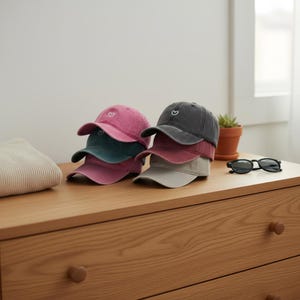 Könnte beinhalten: Mehrere Baseballkappen in Rosa-, Türkis-, Burgunder-, Grau- und Beigetönen sind auf einer Holzoberfläche gestapelt. Jede Kappe hat ein kleines weißes Herz auf der Vorderseite. Eine schwarze Sonnenbrille liegt daneben.