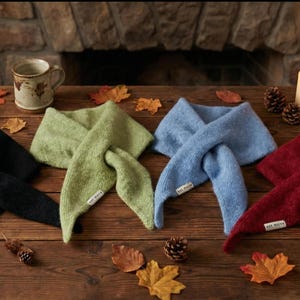 Puede incluir: Varios bufandas de punto en negro, verde, azul y rojo, dispuestas sobre una mesa de madera con hojas de otoño y piñas. Una vela encendida y una taza sugieren un ambiente cálido y otoñal.