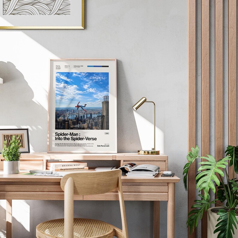 May include: A framed poster for the movie "Spider-Man: Into the Spider-Verse" is displayed on a wooden desk. The poster features a cityscape with Spider-Man falling. A gold desk lamp and a small potted plant are also on the desk.