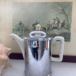 Könnte beinhalten: Eine silberfarbene Vintage-Kaffeekanne mit weißem Keramikgriff und Ausguss. Die Kanne hat eine gehämmerten Textur und einen cremefarbenen Knopf auf dem Deckel. Der Hintergrund zeigt einen gerahmten Druck mit einer historischen Szene.