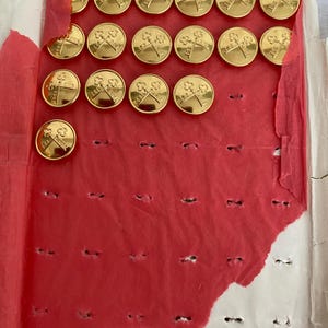 Könnte beinhalten: Eine Reihe goldfarbener Knöpfe mit einem Kreuz- und Schlüsseldesign, angeordnet auf einem roten Papierrücken. Die Knöpfe sind rund und glänzend, mit einem detaillierten Emblem. Das Papier ist stellenweise zerrissen und gibt einen weißen Hintergrund frei.
