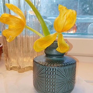 May include: A teal ceramic vase with a geometric pattern holds two yellow tulips. The vase has a round body and a narrow neck. The tulips have bright yellow petals and green stems. The vase is on a white surface.