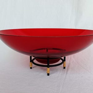 Può includere: Una grande ciotola di vetro rosso rubino con un design ampio e poco profondo. La ciotola poggia su un supporto in metallo nero con quattro gambe, rifinito con materiale intrecciato color beige. Il bordo della ciotola ha un bordo sottile e scuro.