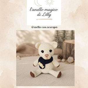 Può includere: Un orsetto all'uncinetto color crema con zampe marrone scuro e una sciarpa blu scuro con un piccolo bottone a forma di caramella. L'immagine include il testo l'anello magico di Lilly, Orsetto con sciarpa e Livello intermedio – PDF pattern in Italiano e Inglese.