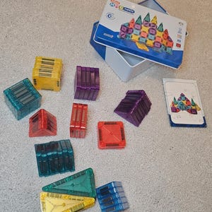 May include: A collection of Mini Magnetic building blocks in assorted shapes and colours, including red, yellow, blue, green, and purple. The set includes a blue and white tin with a picture of a building made from the blocks and a card with building instructions.
