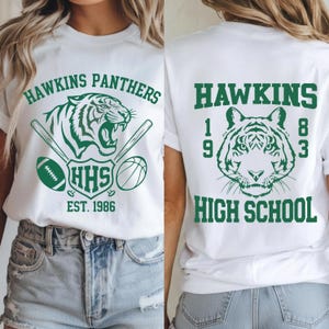 May include: White t-shirt with a green graphic featuring a tiger, baseball bats, a football, and a basketball. The text "HAWKINS PANTHERS" arches above the tiger, with "HHS" and "EST. 1986" below. The back of the shirt displays a tiger and the text "HAWKINS HIGH SCHOOL 1983".